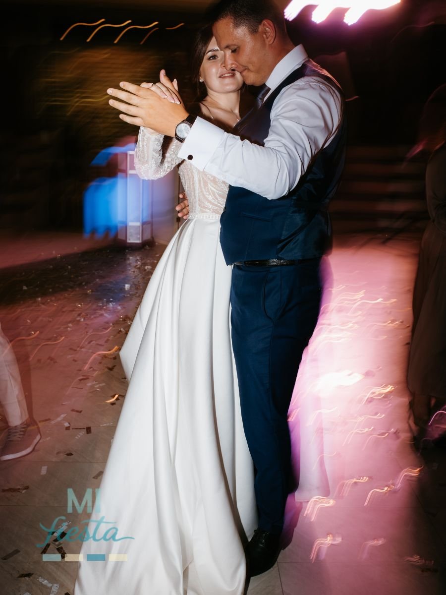 A newlywed couple shares a romantic wedding dance, with the bride in a flowing white dress and the groom in a navy vest and tie.